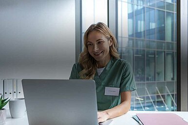 Medizinische Fachkraft in grüner Arbeitskleidung arbeitet am Laptop in modernem Büro mit Panoramafenstern