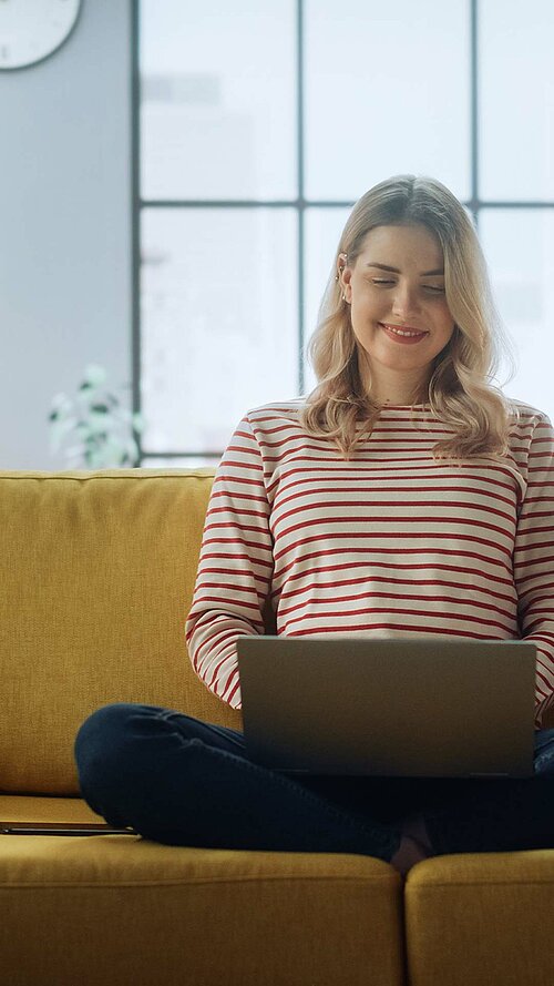 Person sitzt auf einem gelben Sofa und arbeitet am Laptop in einem modernen Wohnzimmer mit großen Fenstern