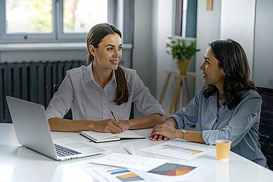 Zwei Geschäftsfrauen bei einem Meeting im modernen Büro, mit Laptop, Notizbuch und Diagrammen auf dem Tisch