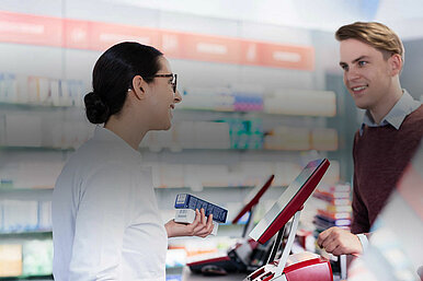Apothekerin berät Kunden an der Kasse einer modernen Apotheke mit Medikamentenregalen im Hintergrund