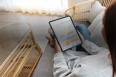 Person sitzt auf einem Sofa und betrachtet Datenanalysen auf einem Tablet, das verschiedene Diagramme und Grafiken anzeigt