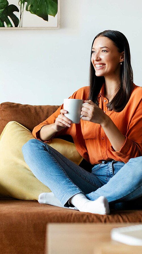 Frau in orangefarbenem Oberteil und blauer Jeans sitzt entspannt auf braunem Sofa mit Tasse in der Hand